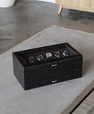 The Bayswater 12 Slot Watch Box by TAWBURY, complete with a transparent lid and black/charcoal finish, showcases five watches on a grey table, ideal for any collector's essentials.