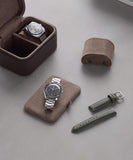 A silver wristwatch rests on a suede pad, paired with a green leather strap and the Fraser 2 Watch Travel Case - Brown by TAWBURY, complete with a plush cushion, all elegantly arranged on a gray surface.
