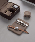 Two watches are elegantly displayed: one in a sleek TAWBURY Fraser 3 Watch Travel Case with a metal strap and tool, while the other, with a brown leather strap, is nestled on a suede pouch beside a soft cushion.