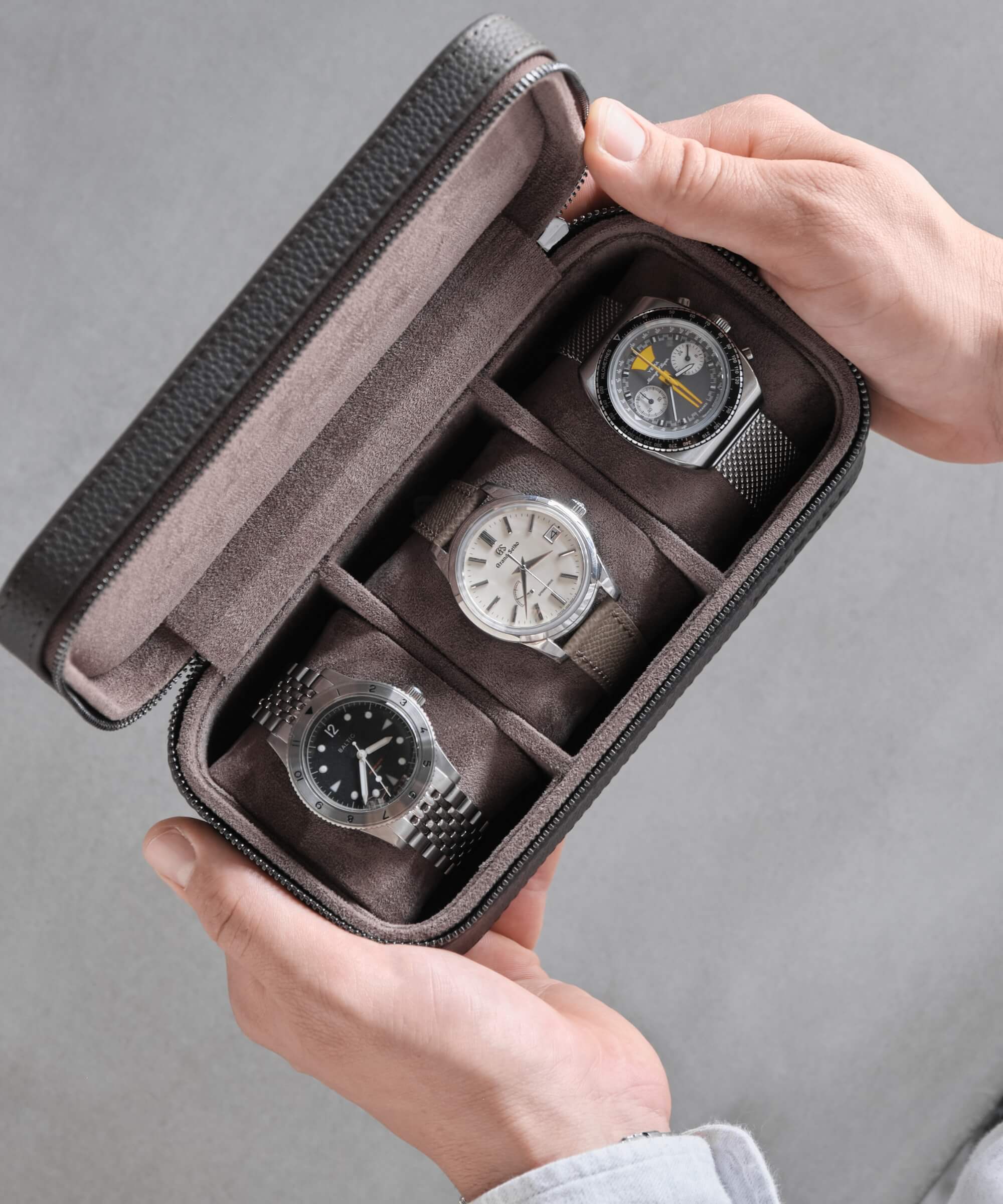 A person holding a Fraser 3 Watch Travel Case - Brown from TAWBURY with three watches in it. The case is made from genuine leather and features a sleek black exterior with red stitching accents. Whether you're traveling for business or pleasure, the