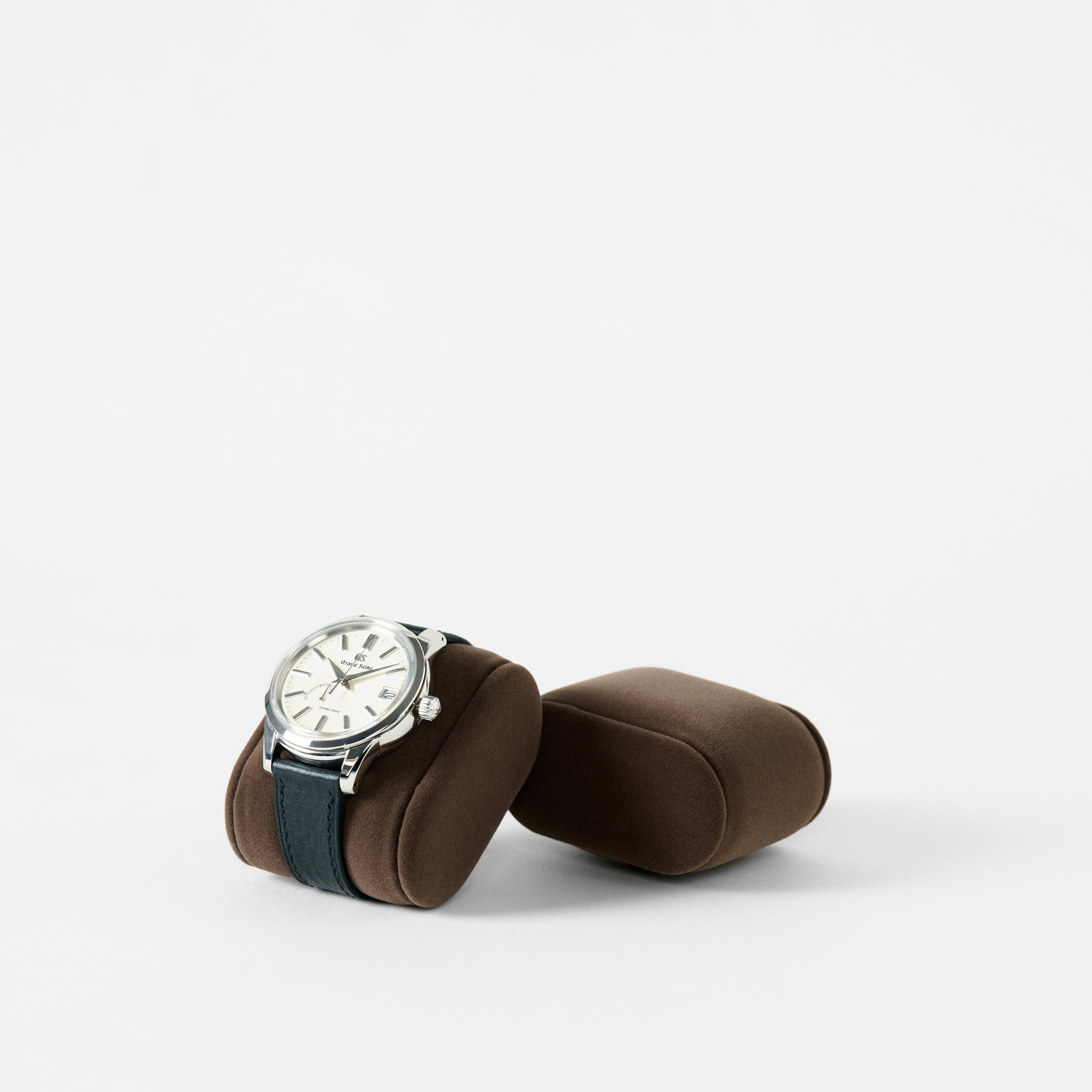 A silver wristwatch with a black band is shown on a TAWBURY Bayswater Watch Pillow (Brown/Grey, 55mm, Small), with another matching pillow beside it, set against a plain white background.