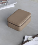 A taupe rectangular leather watch case, the Fraser 4 Watch Travel Case by TAWBURY, sits on a gray surface near a wristwatch, a leather strap, and some open books and magazines.