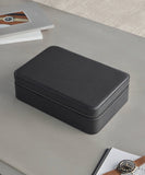 The Fraser 8 Watch Case - Black by TAWBURY, a rectangular leather zippered case, sits on a gray surface with books and a magazine nearby, appealing to discerning watch collectors.