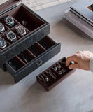 A hand places a small tray with rings and cufflinks beside an open TAWBURY Bayswater 6 Watch Jewellery Box in Black/Charcoal, displaying six wristwatches and accessories.