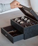 A person opens the TAWBURY Bayswater 6 Watch Jewellery Box in black/charcoal, revealing six watches plus cufflinks, bracelets, and sunglasses organized in a pull-out drawer.