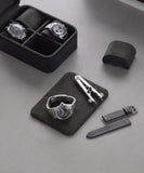 The TAWBURY Fraser 4 Watch Travel Case in black displays exquisite watches on a gray surface, accompanied by two extra strap bands, a silver tool, and another watch presented separately.