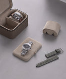 Two watches are elegantly displayed on beige holders, one in a Fraser 2 Watch Travel Case by TAWBURY. A green textured strap and cushion rest nearby, all on a refined gray surface, ensuring exceptional protection.