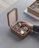 A person is holding a TAWBURY Fraser 4 Watch Travel Case in taupe with four wristwatches inside, while another watch box containing watches is placed on the surface nearby.