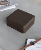 The Fraser 6 Watch Case - Brown by TAWBURY rests on a gray surface, accompanied by a wristwatch, book, and magazine.