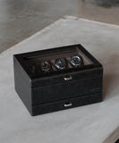 The TAWBURY Bayswater 8 Slot Watch Box in black vegan leather with a glass top elegantly showcases four watches on a light-colored table, perfect for enthusiasts.