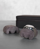 The TAWBURY Fraser Watch Case Pillows in X-Small Brown enhance the display of a silver wristwatch on a gray suede cushion, while an empty slender pillow sits nearby. In the background are watch slots within slightly blurred black leather Fraser Brown Leather Watch Travel Cases.