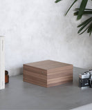 The Grove 6 Slot Wooden Watch Box by TAWBURY, with its exquisite Walnut Wood veneer, complements the wooden box on a gray countertop, surrounded by a book, small jar, vintage camera, and plant in the background.