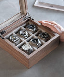 A hand opens a TAWBURY Grove 6 Slot Wooden Watch Box made from Walnut Wood, showcasing six wristwatches with various designs and bands, all neatly arranged in individual compartments beneath the glass lid.