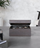 A TAWBURY Grove 6 Slot Wooden Watch Box in Kassod Wood with a solid lid displays four wristwatches on a gray table, against a blurred background of a chair and plant.