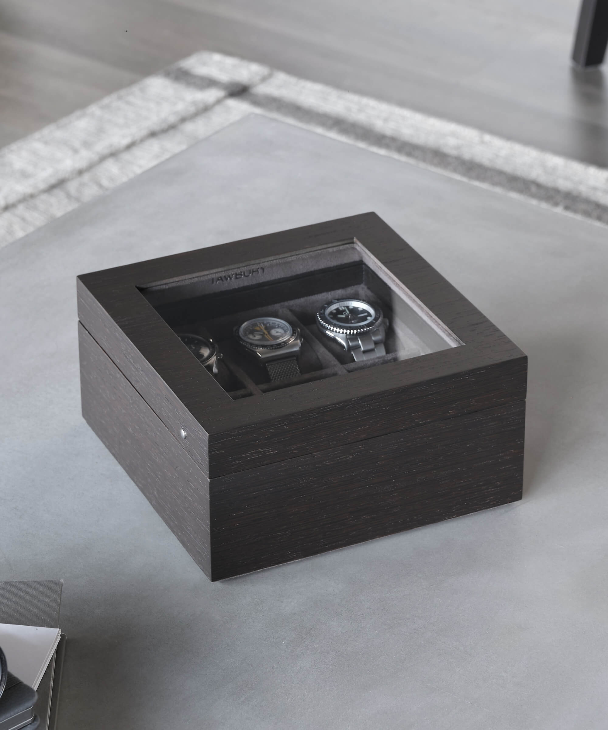 A TAWBURY Grove 6 Slot Watch Box with Glass Lid - Kassod on a table with two timepieces in it.