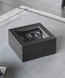 A Grove 6 Slot Wooden Watch Box by TAWBURY, crafted from elegant Kassod wood veneer and featuring a glass lid, displays three wristwatches inside and is placed on a table.