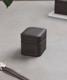 The TAWBURY Fraser 1 Watch Travel Case in brown leather rests on a light gray surface next to a wristwatch with a matching brown leather band.