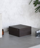 A TAWBURY Grove 6 Slot Wooden Watch Box, made from Kassod Wood and featuring a solid lid, sits on a grey surface next to a book, a candle, and a vintage camera with palm leaves in the background.