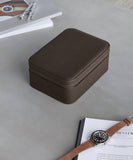 A closed Fraser 4 Watch Travel Case - Brown by TAWBURY sits on a gray surface next to a wristwatch, an open book, and a magazine.
