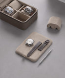 A TAWBURY Fraser 4 Watch Travel Case in taupe rests on a grey surface, holding interchangeable straps, a dual-ended tool, and two elegant watches.