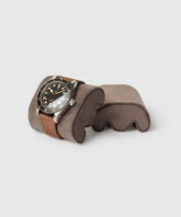 The TAWBURY Fraser Watch Case Pillow in X-Small, Brown elegantly displays a wristwatch with a black dial and silver rotating bezel on its slender form.