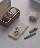 Two wristwatches from the Fraser Collection are placed in a TAWBURY Fraser 3 Watch Travel Case - Taupe with extra straps on a gray surface, alongside an additional watch cushion.