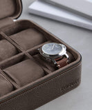 The Fraser 8 Watch Case by TAWBURY cradles a brown leather watch, elegantly set on a light gray surface with closed books in the backgroundāa perfect scene for discerning watch collectors.