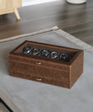 The Bayswater 12 Slot Watch Box with Storage in brown by TAWBURY, featuring a glass top that beautifully displays six wristwatches, is placed on a light-colored surface in a room with wooden flooring, making it an ideal choice for watch enthusiasts.