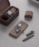 Two wristwatches are elegantly displayed: one in a Fraser 3 Watch Travel Case - Brown and the other on a cushion. Nearby, a green strap with matching buckle lies on the sleek gray surface, showcasing TAWBURY's refined style in leather watch cases.