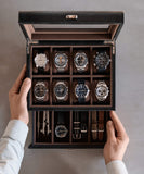 A person showcases the Bayswater 8 Slot Watch Box with Storage in Black/Grey by TAWBURY, presenting eight luxury watches and five extra straps on a gray surface. Ideal for watch enthusiasts who value elegant vegan leather accessories.