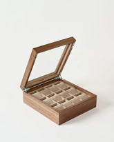 The TAWBURY Grove 12 Slot Wooden Watch Box in light walnut features a glass lid and multiple beige velvet-lined compartments, perfect for organizing your watches or jewelry.