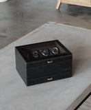 The Bayswater 8 Slot Watch Box by TAWBURY features a black/charcoal finish and a glass lid, offering an elegant way to display three watches on a gray tabletop—ideal for any watch collector.