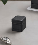 The TAWBURY Fraser 1 Watch Travel Case - Black, featuring textured leather and a zipper, rests on a gray surface beside a silver wristwatch.