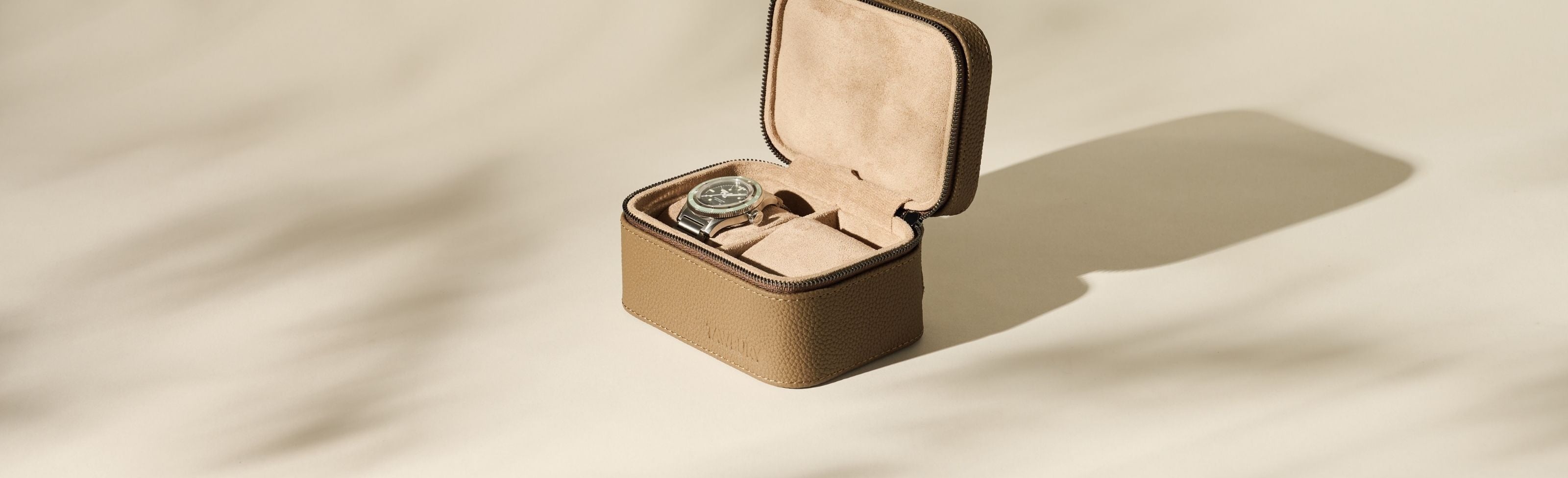 A wristwatch in a brown leather travel case with a suede interior, positioned on a light beige surface with soft shadows.