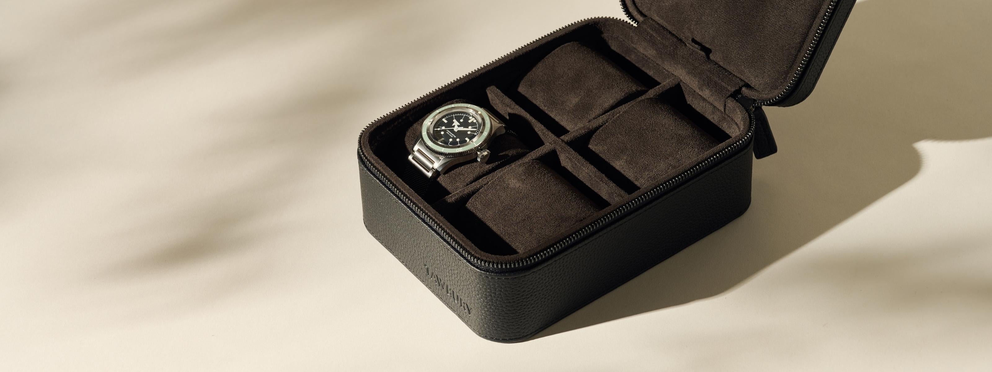 A black leather watch case with a zipper is open, displaying a single wristwatch inside and several empty compartments lined with dark suede.
