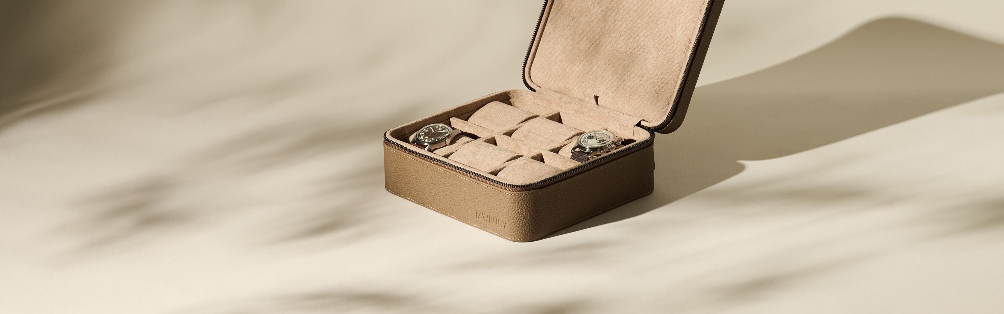 A brown leather watch case with a zipper is open, displaying two wristwatches inside and several empty compartments on a beige surface.