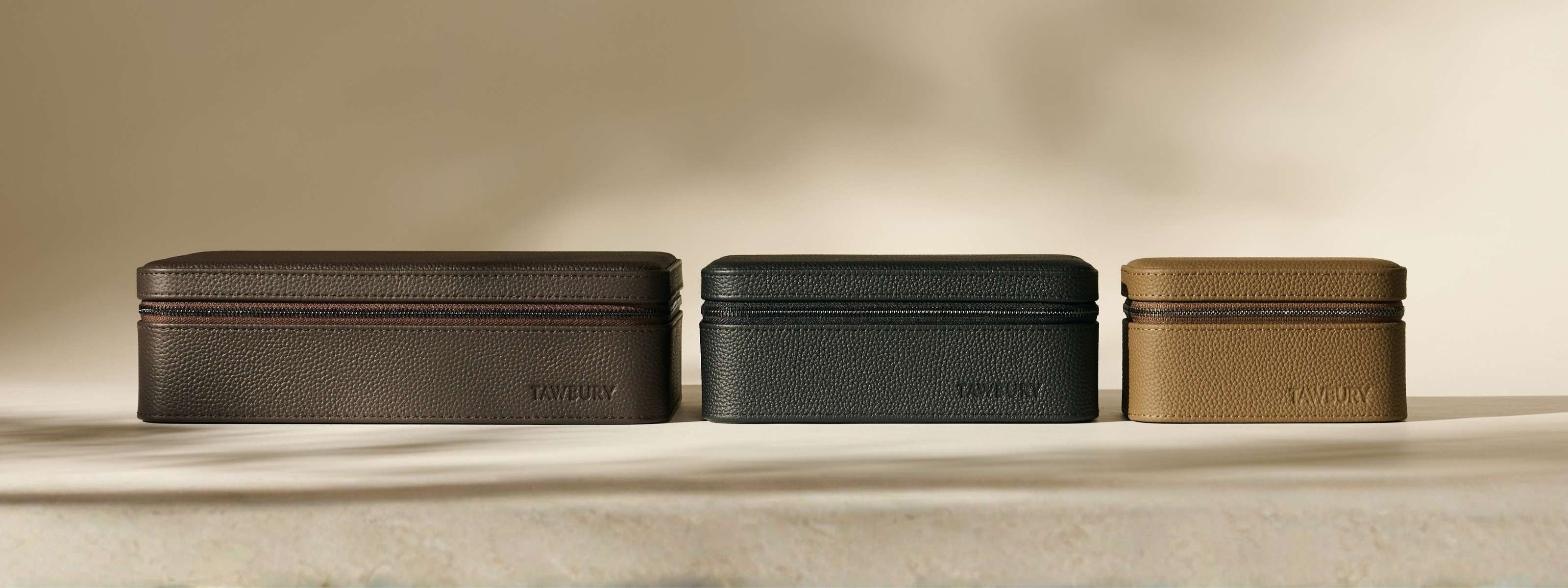 Three textured rectangular leather cases in brown, dark green, and tan are displayed side by side on a neutral surface against a beige background.