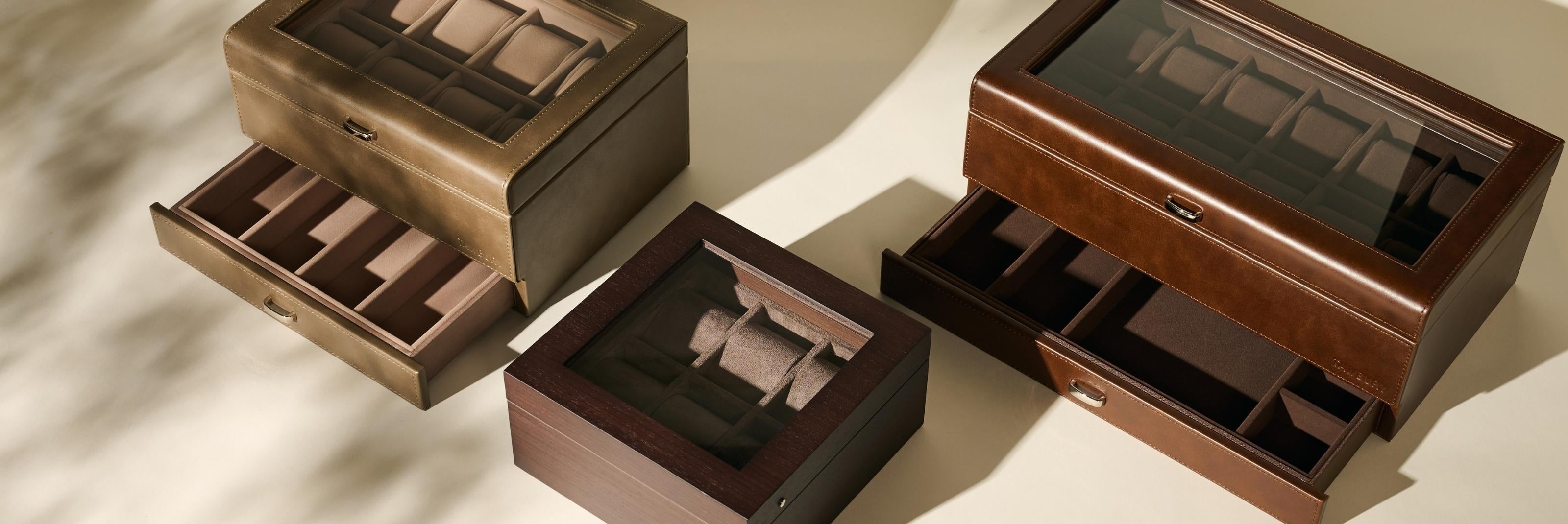 Three glass-topped leather watch boxes in brown and tan are shown, each with multiple compartments for storing watches, with one box having a drawer partially open.
