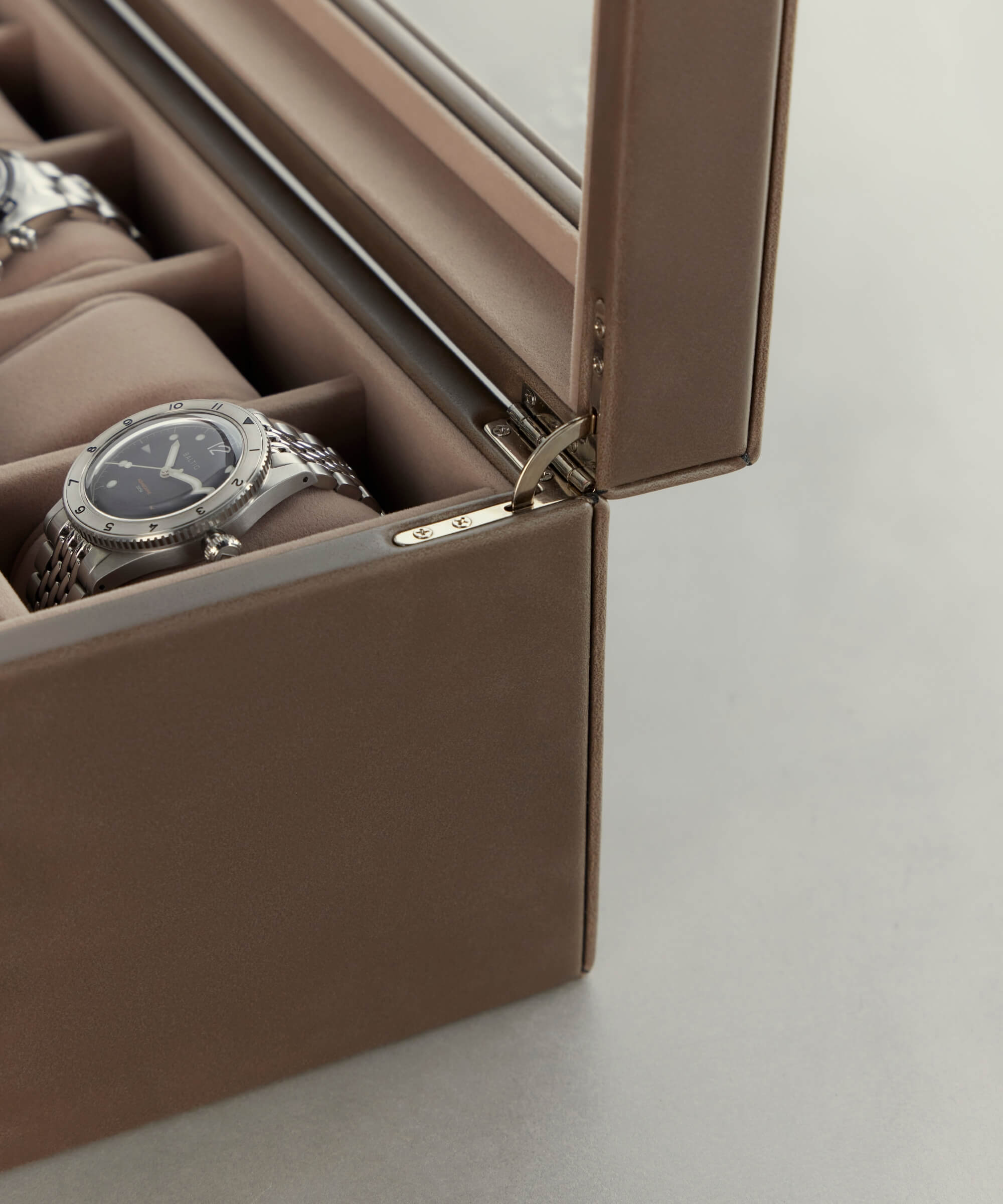 A close-up of the TAWBURY Bayswater 12 Slot Watch Box with Storage in Taupe, crafted from vegan leather with a glass lid, partially open to reveal silver watches inside on a light background—a true treasure for any watch collector.