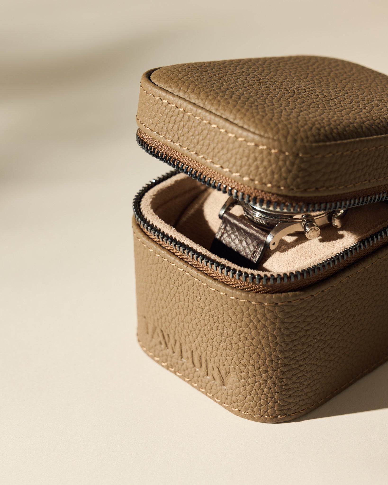 The TAWBURY Fraser 1 Watch Case in taupe, partially unzipped on a light beige surface, reveals a wristwatch inside.