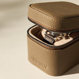 The TAWBURY Fraser 1 Watch Case in taupe, partially unzipped on a light beige surface, reveals a wristwatch inside.