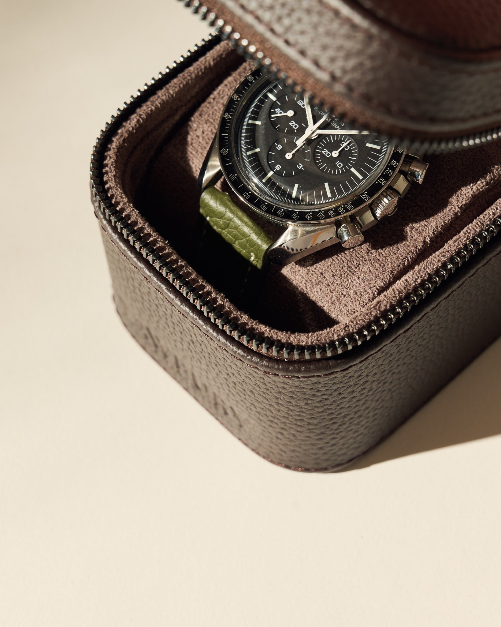 A black chronograph watch with a green strap is kept inside the TAWBURY Fraser 1 Watch Case - Brown, which features textured leather and a soft fabric lining.