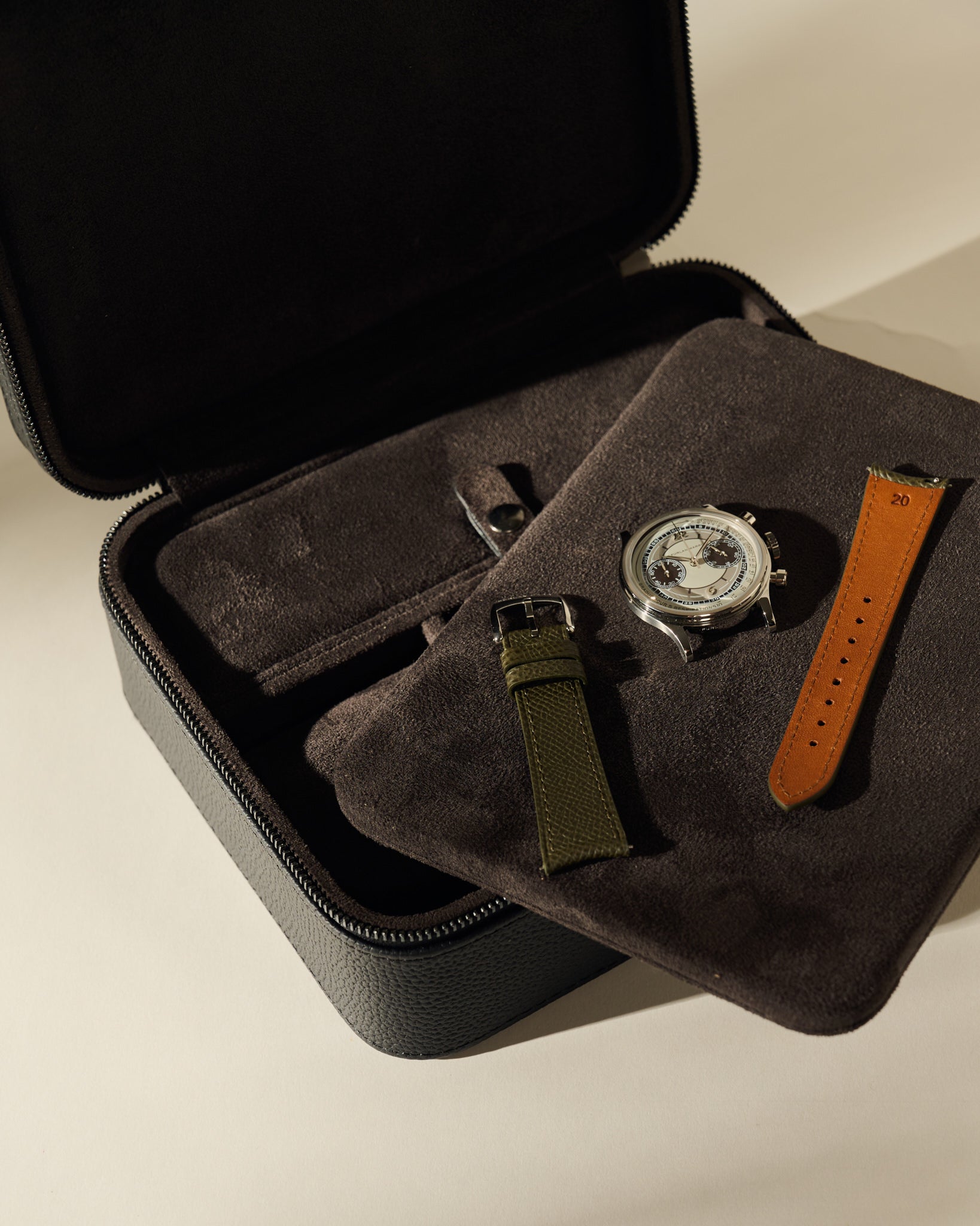 An open TAWBURY Fraser 3 Watch Case with Storage - Midnight Black containing a watch face, a green watch strap, and a brown watch strap resting on a dark fabric divider.