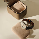 An open TAWBURY Fraser 1 Watch Case - Truffle Taupe with its removable cushion sitting in front, holding a silver wristwatch with a black leather strap.