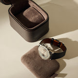 An open, dark brown Fraser 1 Watch Case - Espresso Brown sits behind a luxury wristwatch resting on a matching brown cushion.
