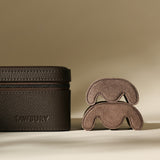 The TAWBURY Fraser 4 Watch Case in Espresso Brown, crafted in textured leather with a zipper, sits beside two curved suede pieces—a stylish and practical case for storing or traveling with up to 4 watches.