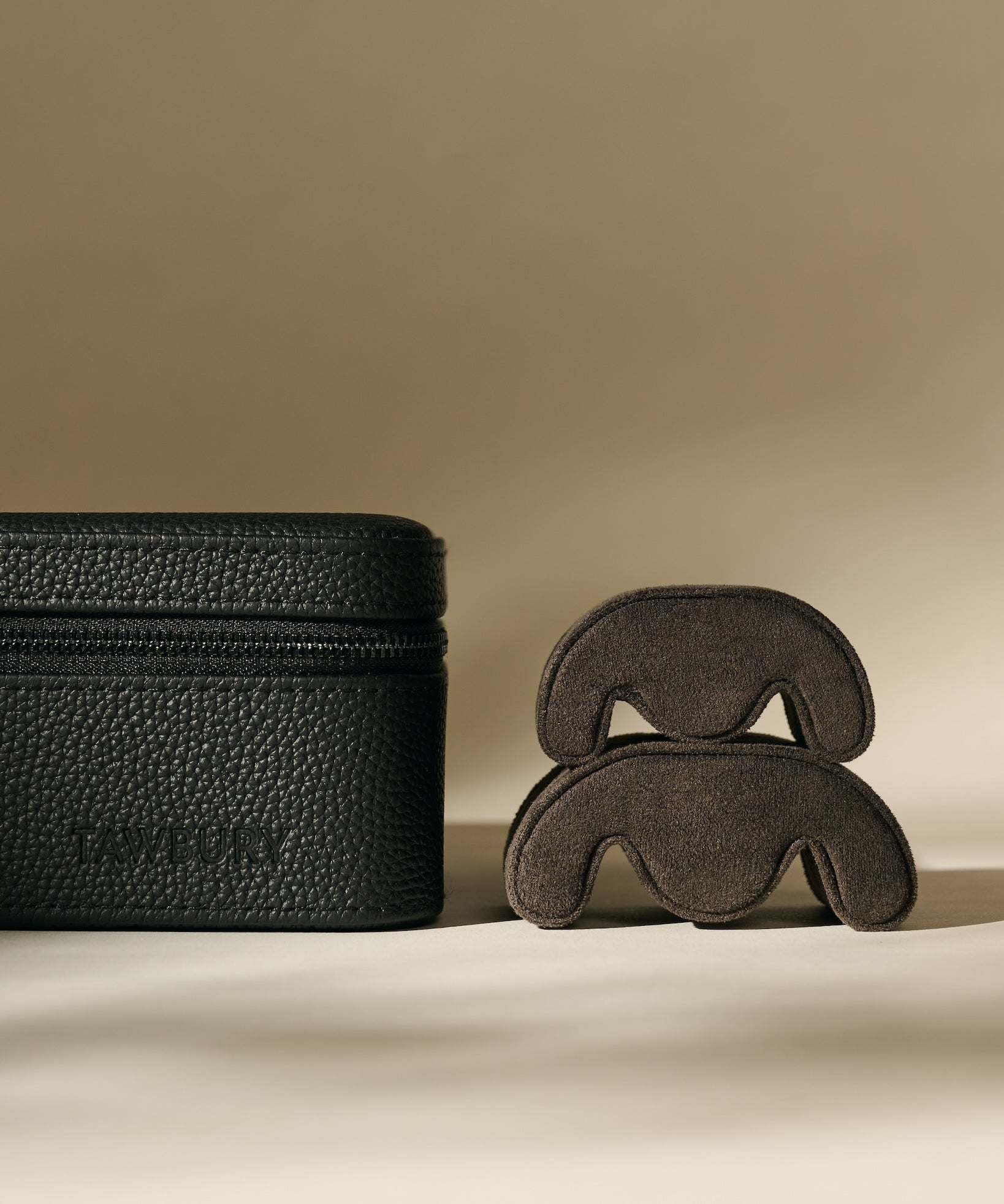 The TAWBURY Fraser 1 Watch Case in Midnight Black is displayed next to two stacked, curved brown suede items on a neutral background.