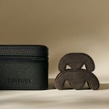The TAWBURY Fraser 6 Watch Case in Midnight Black, featuring a textured leather finish, is displayed beside two curved brown objects on a neutral background.