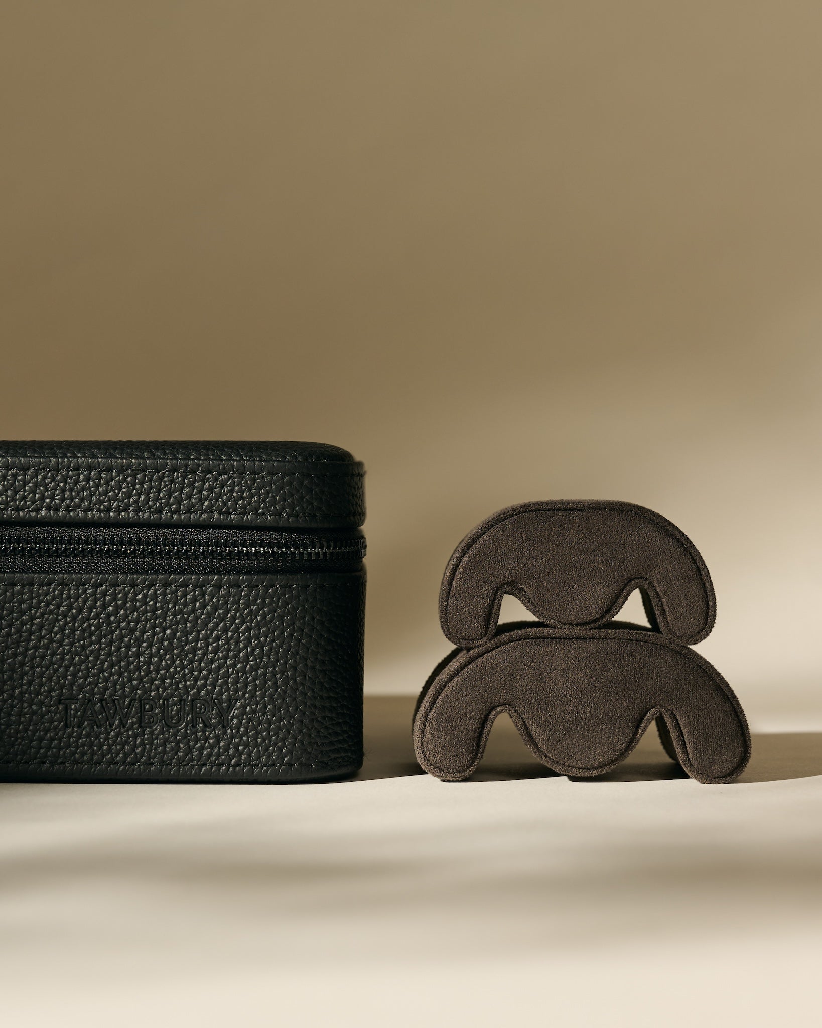 The TAWBURY Fraser 6 Watch Case in Midnight Black, featuring a textured leather finish, is displayed beside two curved brown objects on a neutral background.
