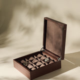 The TAWBURY Grove 12 Slot Wooden Watch Box in dark walnut wood with a solid lid showcases multiple compartments, elegantly organizing two wristwatches and a bracelet on a beige surface with soft shadows.